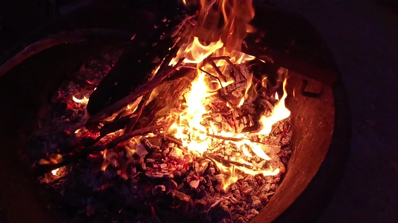 Flames curl across charred wood and ash inside a fire bowl.