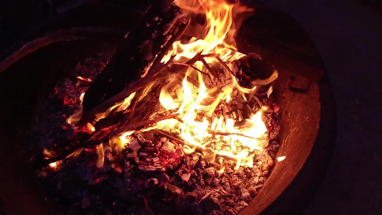 At 0:02, a taller flame flares on the right side of the fire pit while smaller branches and glowing coals remain visible below, with no visible people.