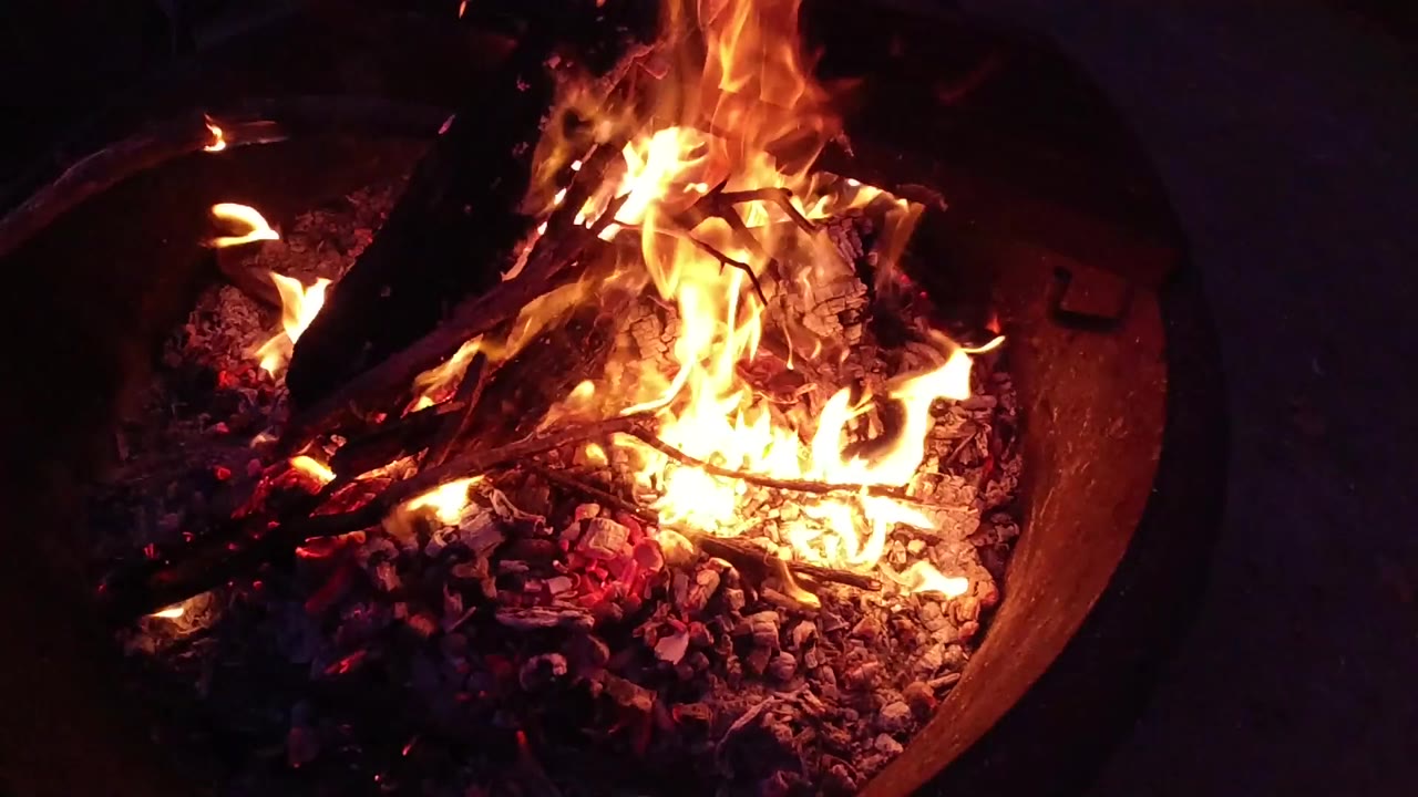 Round metal fire bowl at night with orange flames and glowing embers, viewed from slightly above.