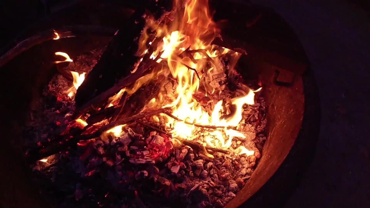 Close-up view of a round metal fire bowl with orange and yellow flames extending well above the rim and glowing embers throughout the base, shot at night against a dark background; no people are visible.