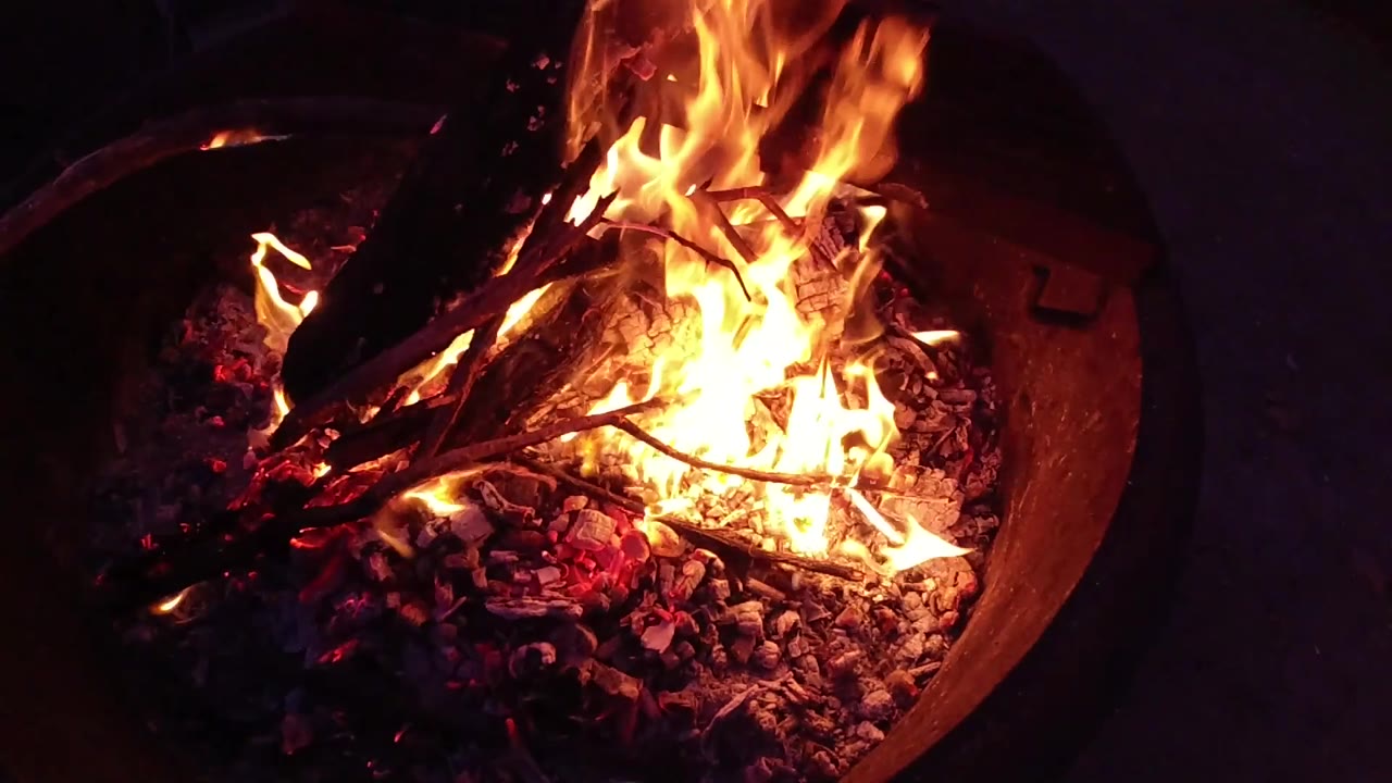 Slightly overhead close-up of a round metal fire bowl with burning wood logs distinguishable among orange flames and glowing embers, the curved metal rim visible at the lower sides of the frame; no people are visible.