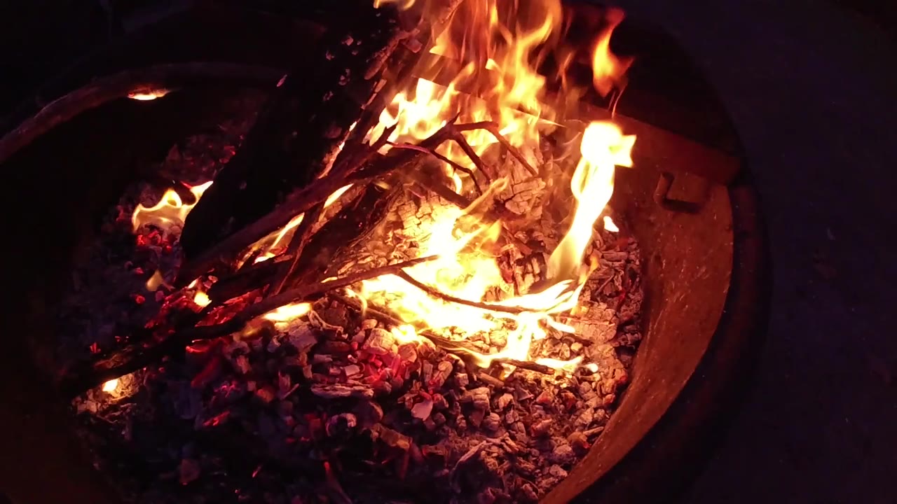 Close-up of a round metal fire bowl at night with a prominent orange flame rising on the left side and bright red-orange embers covering the base, the bowl rim visible at the bottom edge; no people are visible.