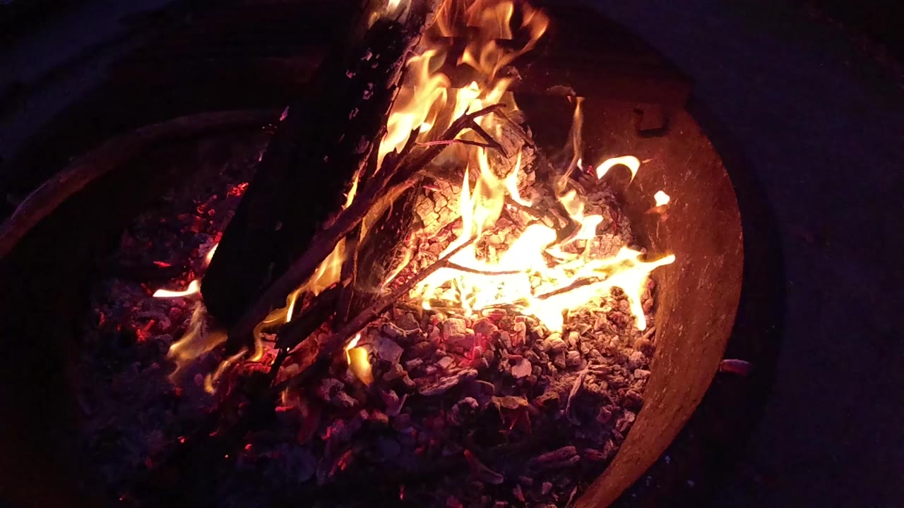 Overhead close-up of a round metal fire barrel at Trestles Beach, California; orange and yellow flames burn around sticks and logs over glowing embers; no visible people