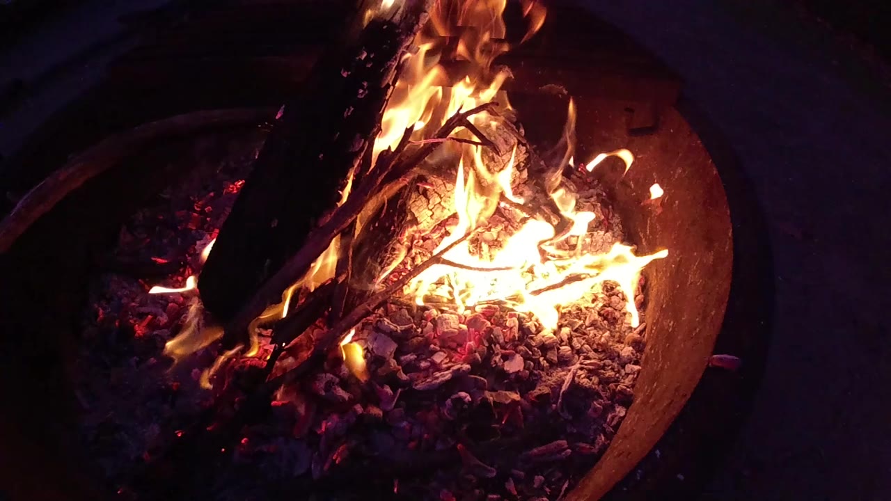 Round metal fire barrel with orange flames and burning sticks visible from above, dark surroundings