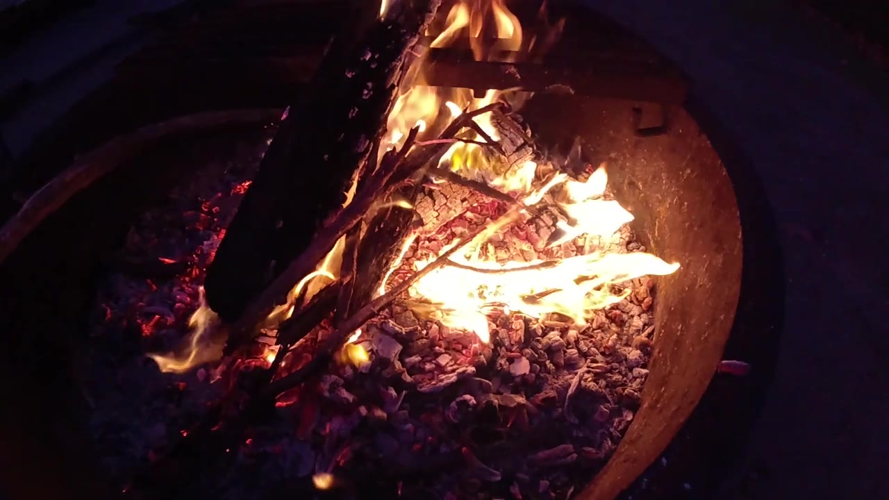 Overhead close-up of a rust-colored round metal fire barrel at Trestles Beach, California; flames and burning logs cast a warm orange glow; no visible people