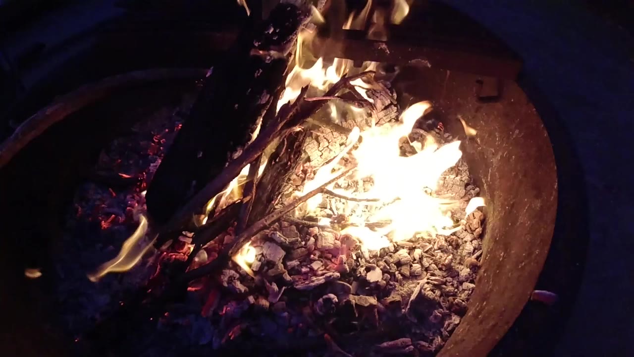 At Trestles Beach, California, a high-angle close-up shows burning wood, pale gravel, ash, and bright flame in a round fire pit with no visible people.