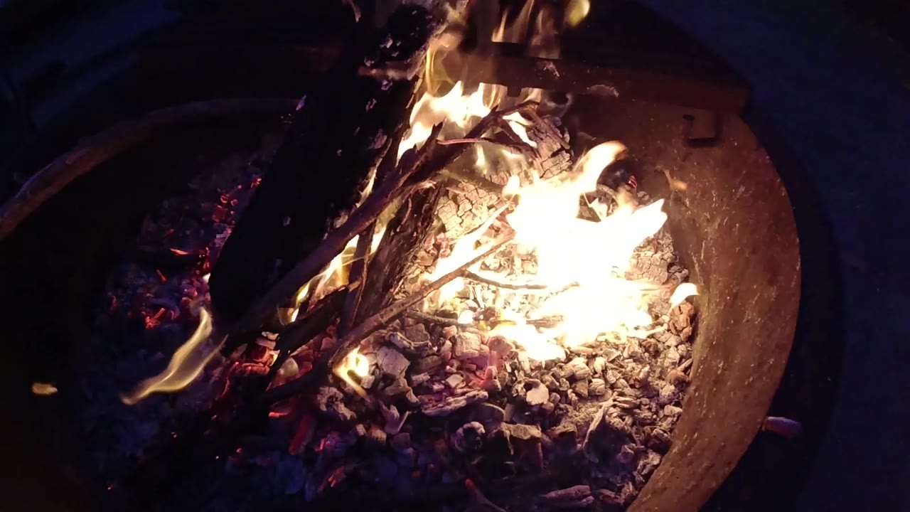Flames flare over ash and gravel beneath charred wood