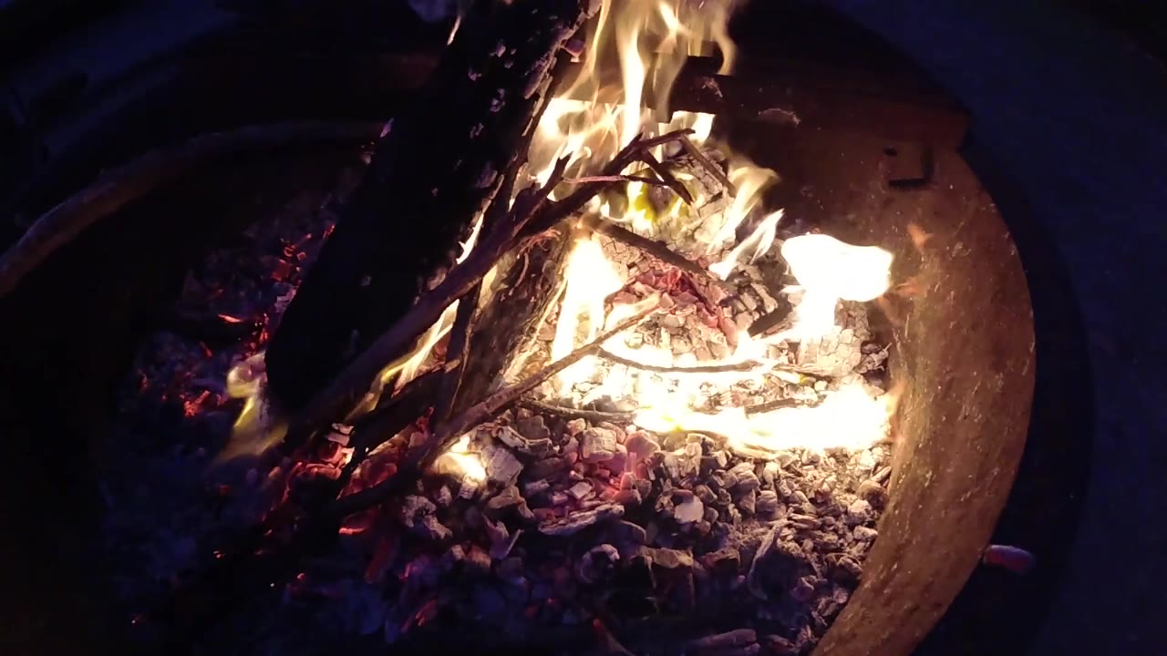 At Trestles Beach, California, a bright tongue of flame rises around crossed sticks and ash inside the round fire pit, with no visible people.