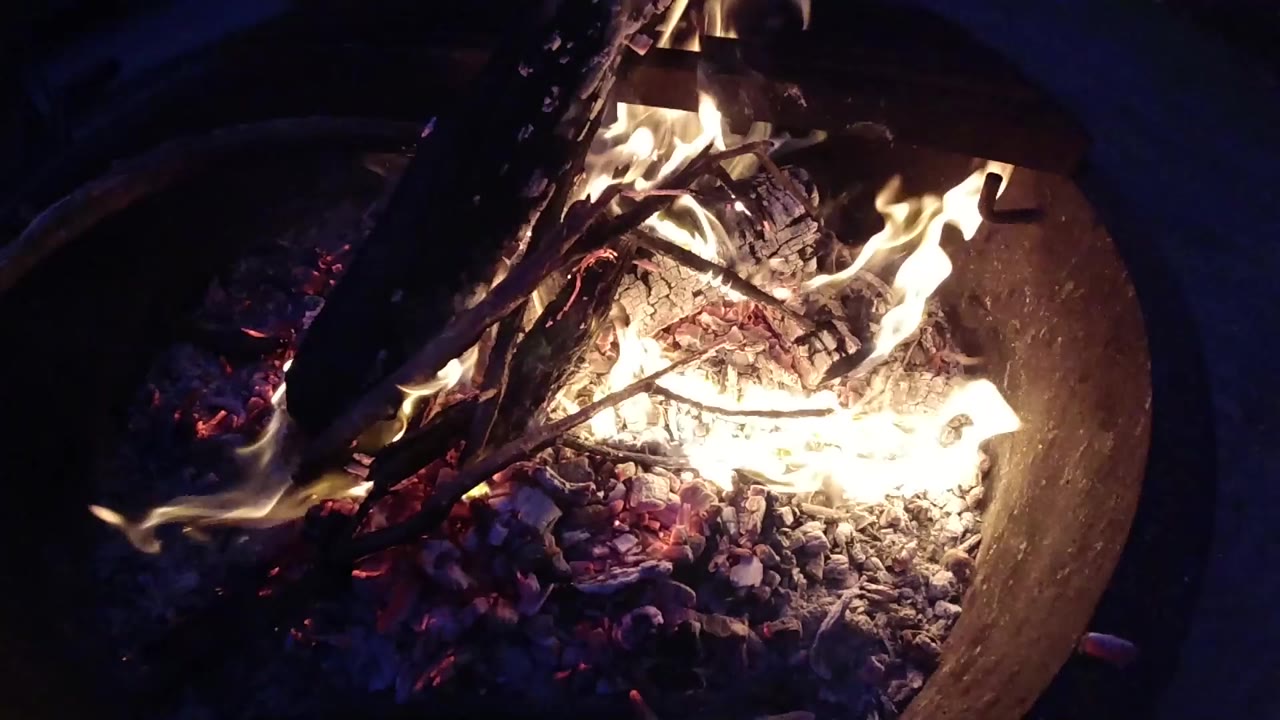 Overhead view of a round metal fire pit at Trestles Beach, California, with burning wood, bright yellow-orange flames, red-orange glowing embers, and ash; no people visible.