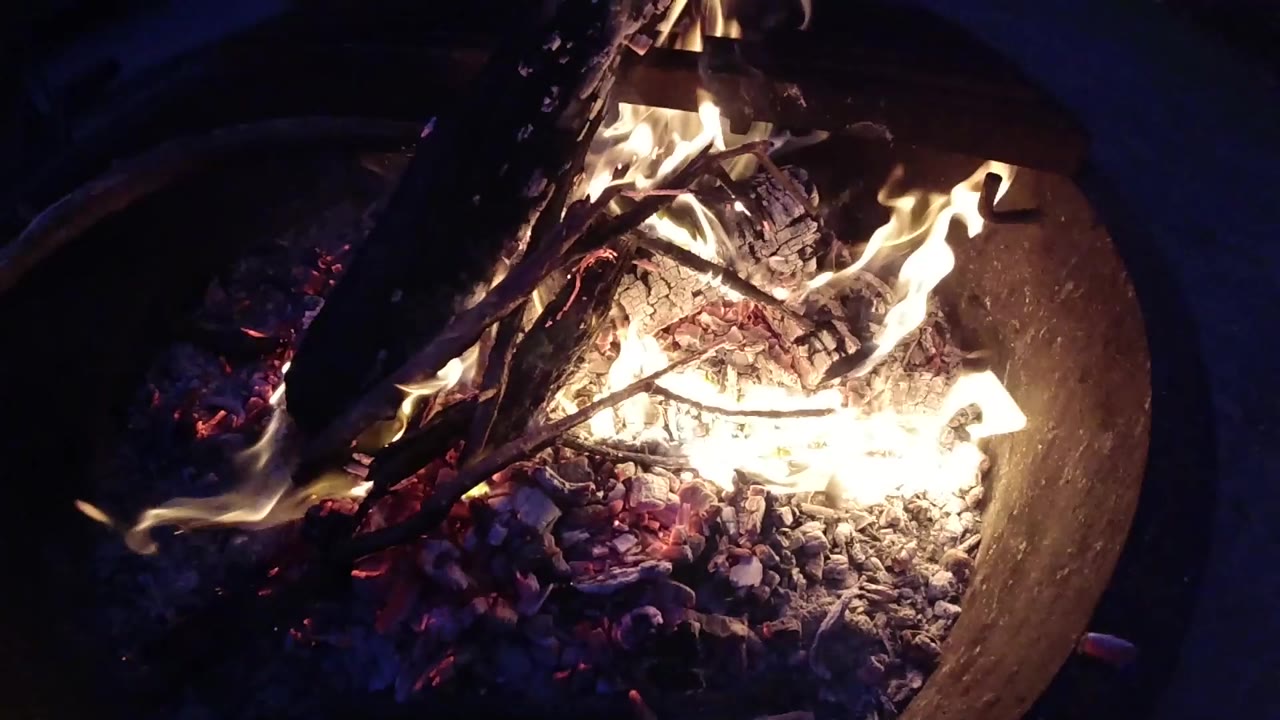 Overhead view of a round metal fire pit at Trestles Beach, California, showing active flames, charred wood, and a glowing ember bed against a dark night background; no people visible.