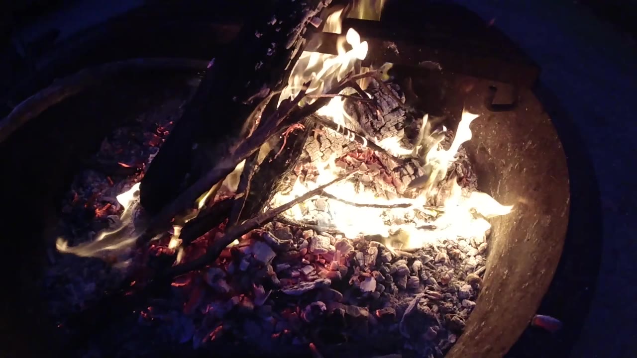 Looking down into a round metal fire pit at Trestles Beach, California, at night, with burning wood, active flames in the upper area, and a bright ember bed; no people visible.