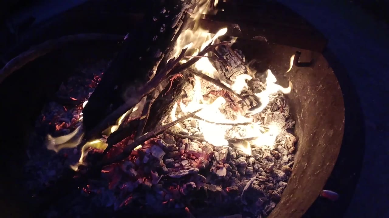 Overhead view of a round metal fire pit at Trestles Beach, California, with active burning wood, yellow-orange flames, and a bright red-orange ember bed; no people visible.
