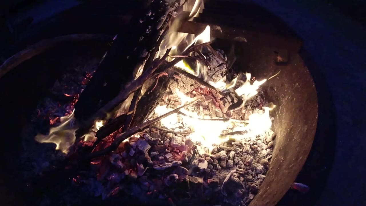 One second into the clip, the overhead view of the metal fire pit at Trestles Beach shows flames redistributed slightly toward the center of the bowl interior as the fire moves naturally, with embers still spread broadly beneath; no people visible.