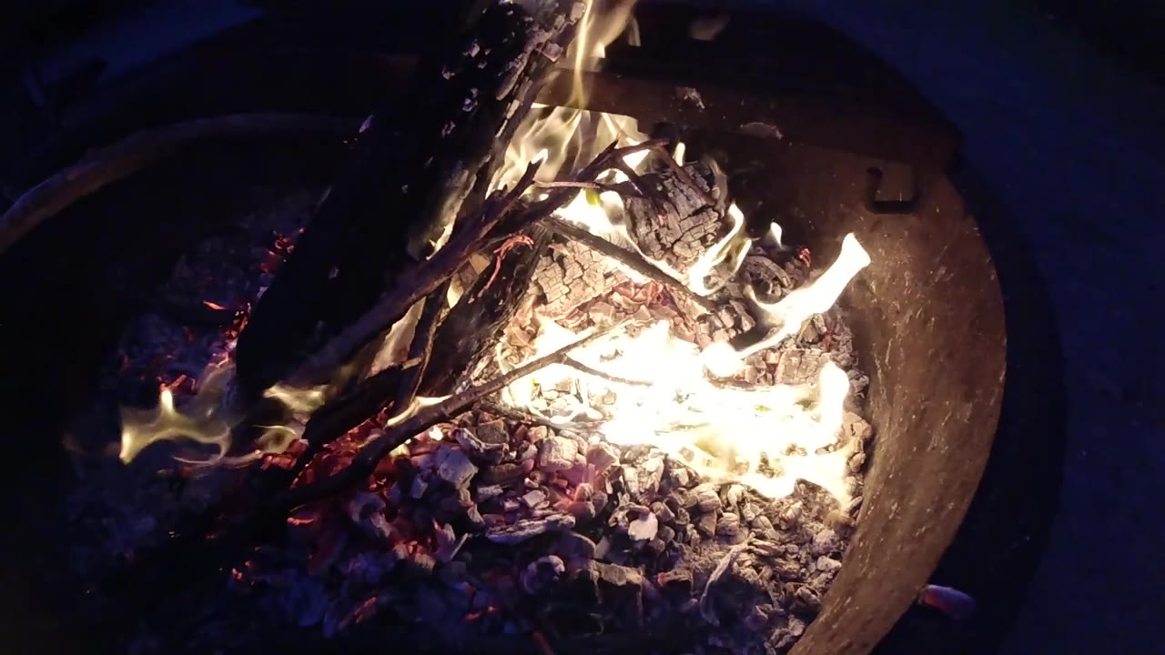 Two seconds in, the drone camera has drifted enough to shift the round metal fire pit toward the right side of the frame, with flames and embers still clearly burning inside the bowl and dark ground filling the left portion; no people visible.