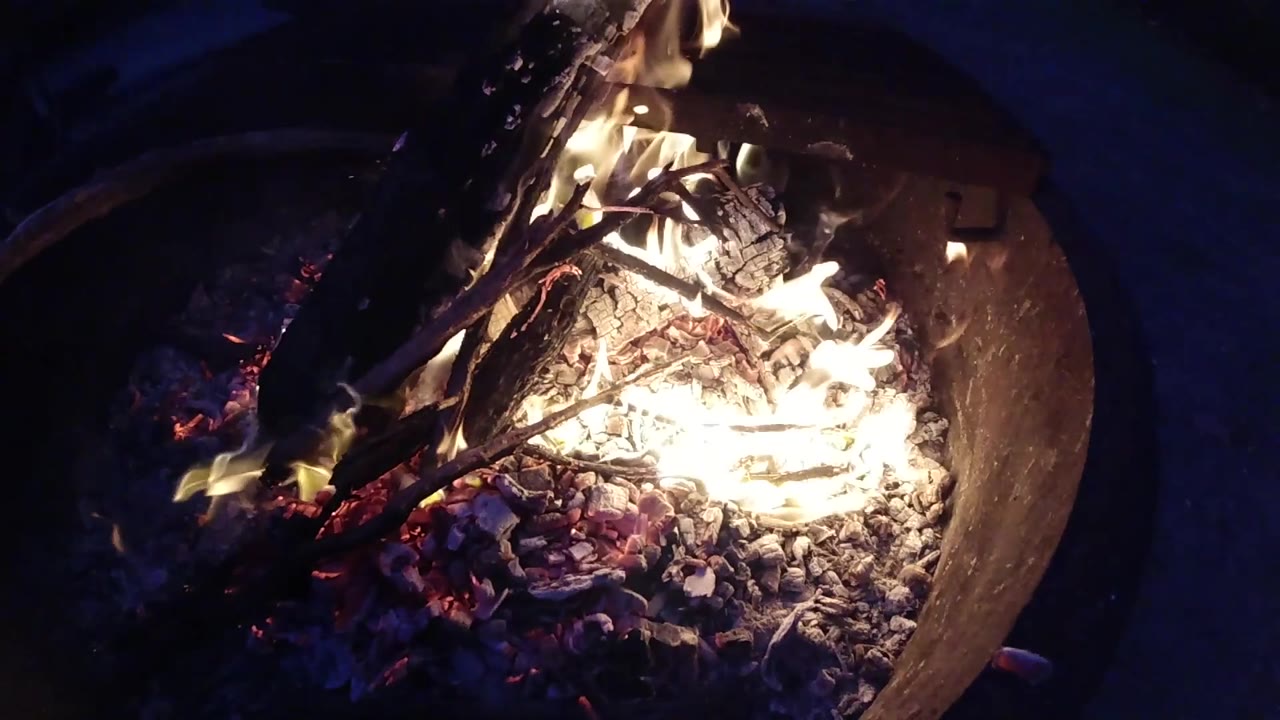 At the midpoint, the drone has repositioned so the metal fire pit now occupies the lower-center of the frame with the bowl rim visible at left, flames burning prominently along the left interior wall and embers glowing across the pit floor; no people visible.