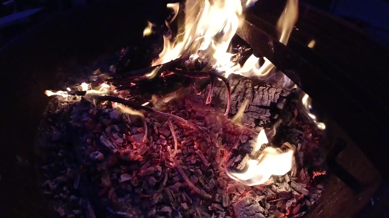 Opening frame of a tight close-up shot of a burning log pile at night at Trestles Beach, California; yellow-orange flames reach upward at the right, a deep red ember bed fills the center, dark surroundings, no people visible.