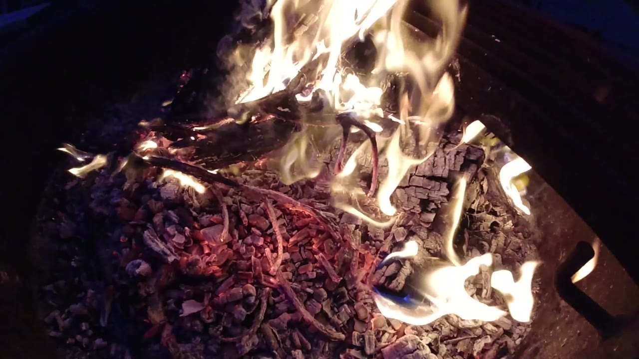 At 0:01 the flame column has shifted slightly toward the upper-right compared to the opening frame, while the red and purple ember bed remains fully active beneath; no people visible, dark surroundings.