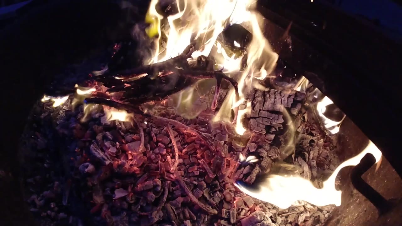 At 0:02 the fire shows distinct flame activity on both the left and right edges of the burning material, with the ember-filled center visible between them; dark fire structure edges appear in the corners and no people are visible.