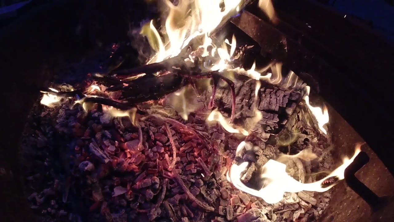 At 0:03 a taller orange flame rises from the upper-left of the fire bed, the most visually dominant flame in the clip, while red and purple embers glow across the lower portion; no people visible.