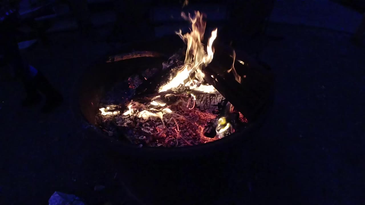 Campfire in a metal fire ring at night, tall yellow-orange flames rising above glowing red-orange embers, shot from an elevated downward angle at Trestles Beach California; no visible people