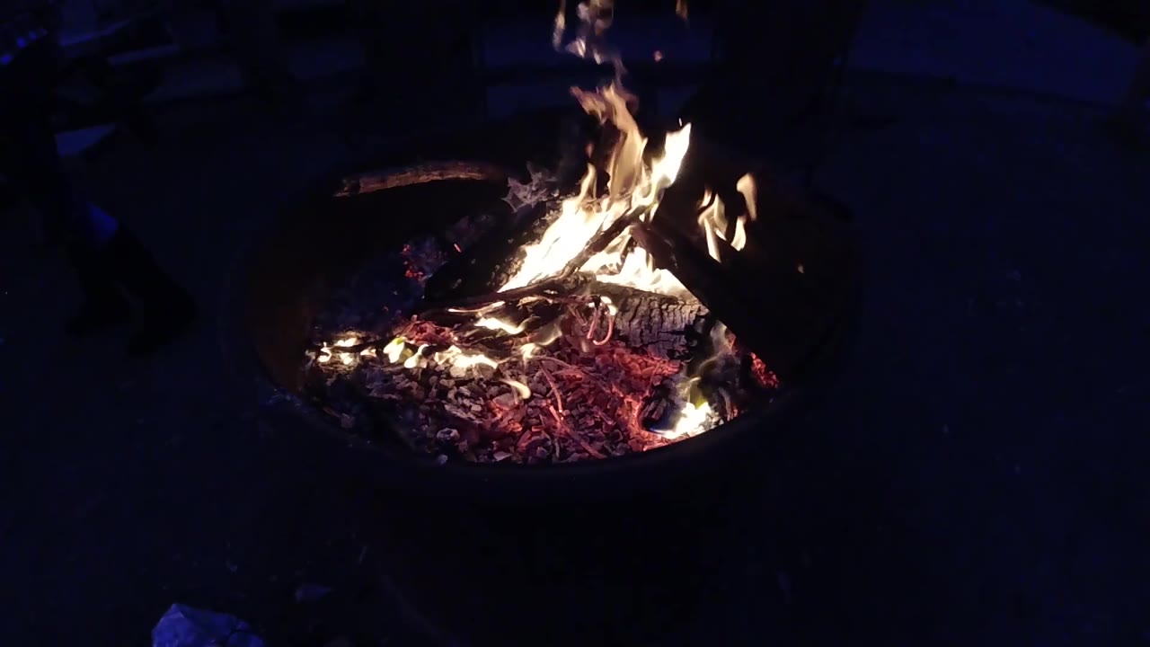 Campfire in a metal fire ring at night, flames tapering to a narrower form compared to the prior second, orange glow visible across the embers; no visible people