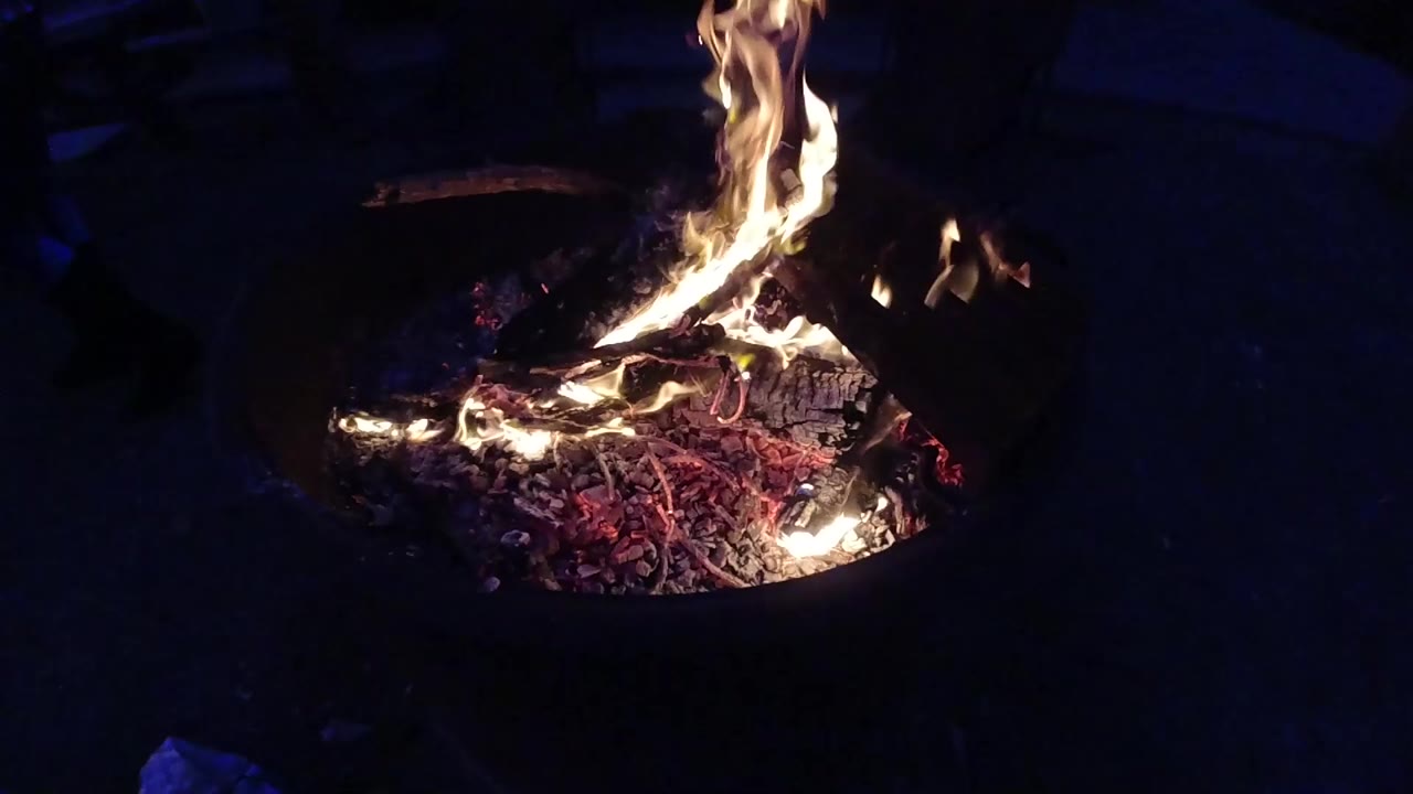 Medium-distance view of a round dark metal fire container at night, with orange and yellow flames rising above it and red embers glowing inside; no visible people and no beach geography identifiable