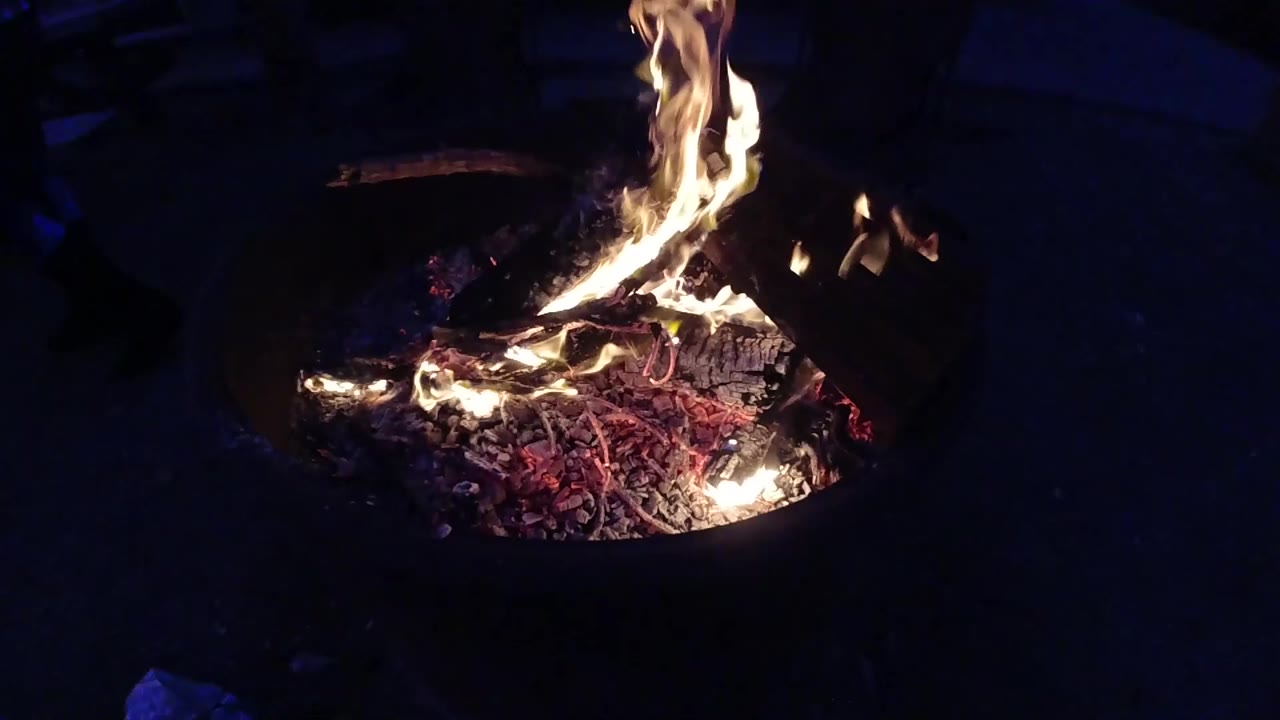 Second frame of the metal fire container at night; flames have shifted slightly in shape from the opening frame while the red embers and burning debris remain consistent; no people visible