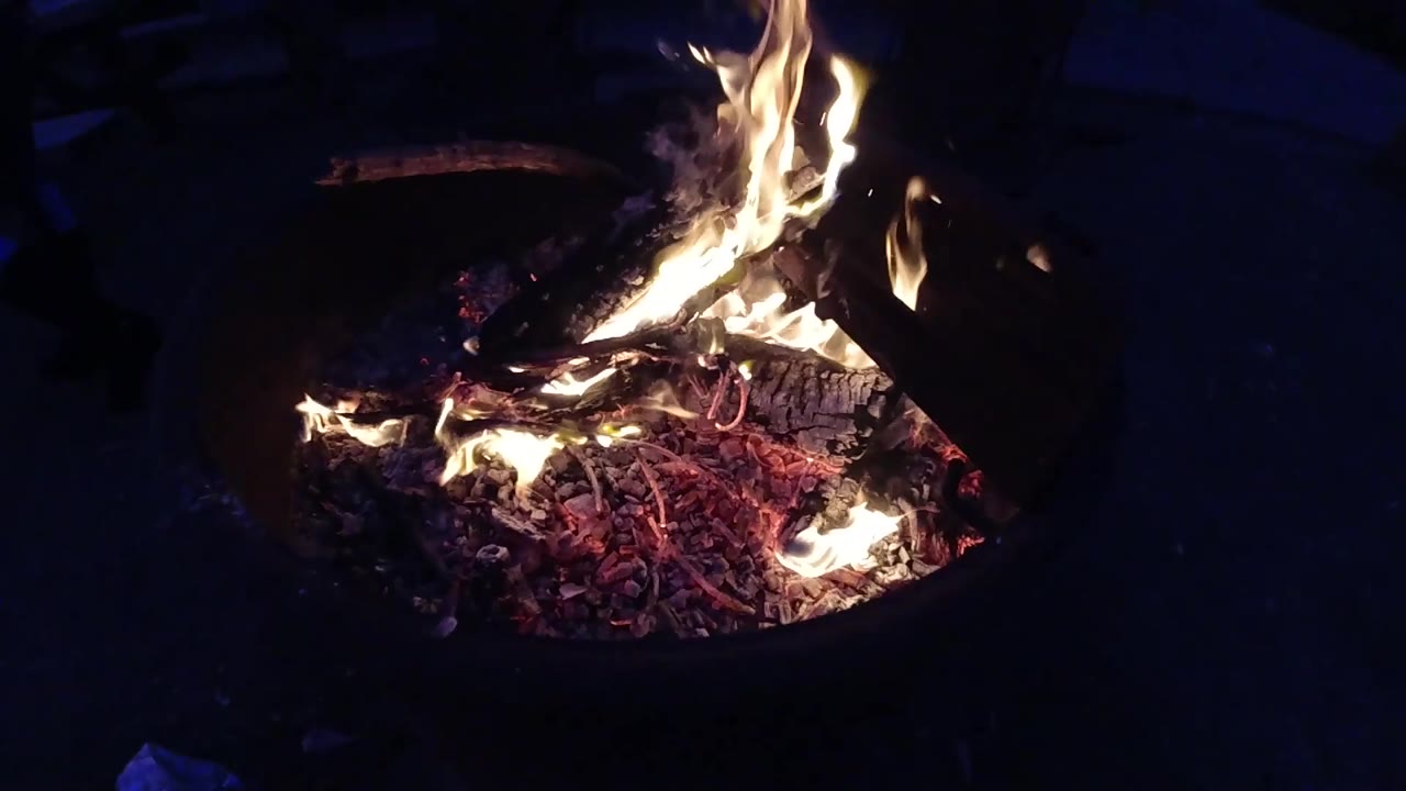 Third frame showing the metal fire container from a similar medium distance; flame shape has changed again from the prior frame, with the dark surroundings remaining consistent throughout; no people are visible