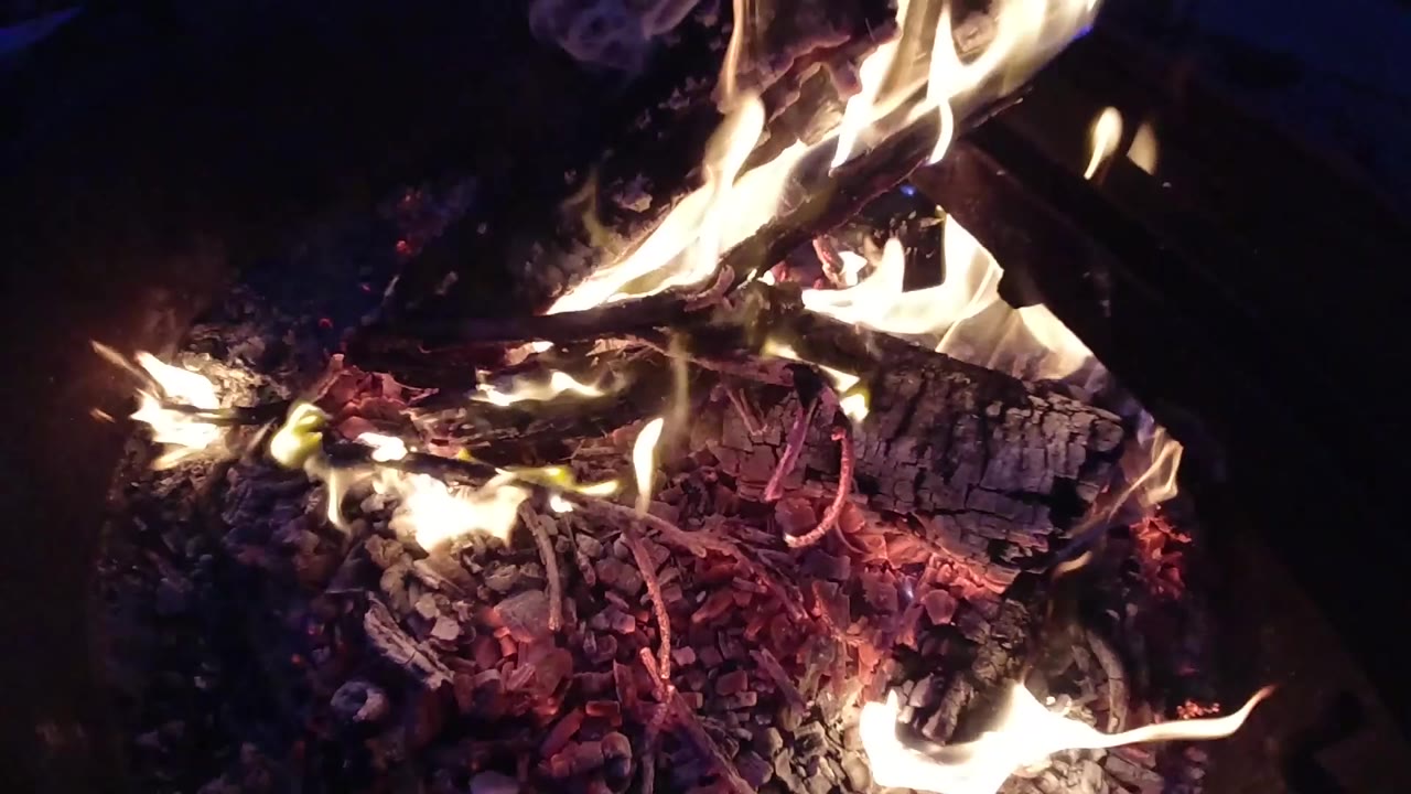 Tight overhead drone shot of a campfire burning in a fire ring at night; large orange and yellow flames fill the upper frame over a bed of red embers; no people visible