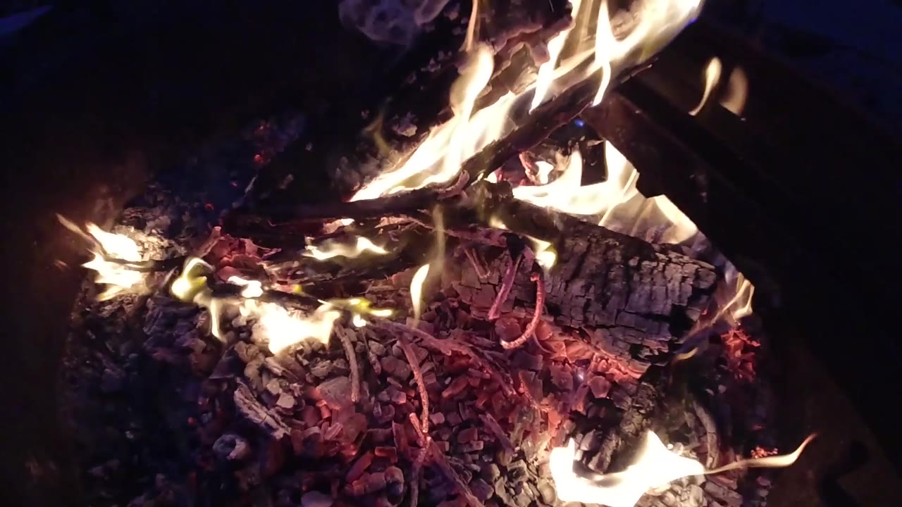 Overhead shot of the campfire at one second, with the flame mass spreading wider and lower compared to frame zero; embers glow beneath; no people visible