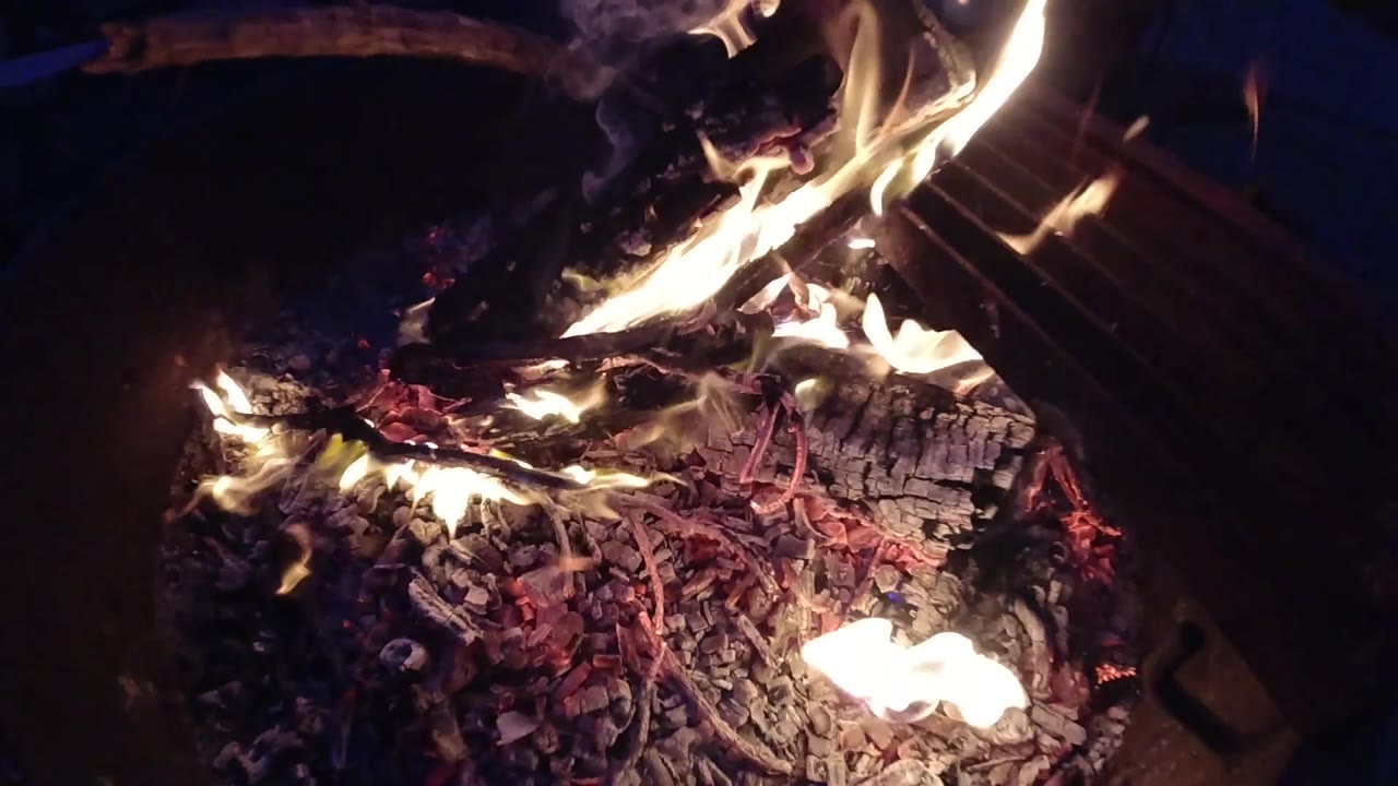Overhead campfire at two seconds, with the flame column leaning and the ember bed more exposed than in the first two frames; no people visible