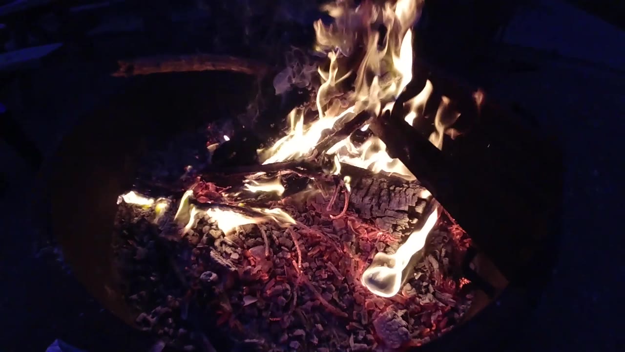 Campfire in a circular metal fire ring with the camera pulled back to show the full pit and dark surrounding ground