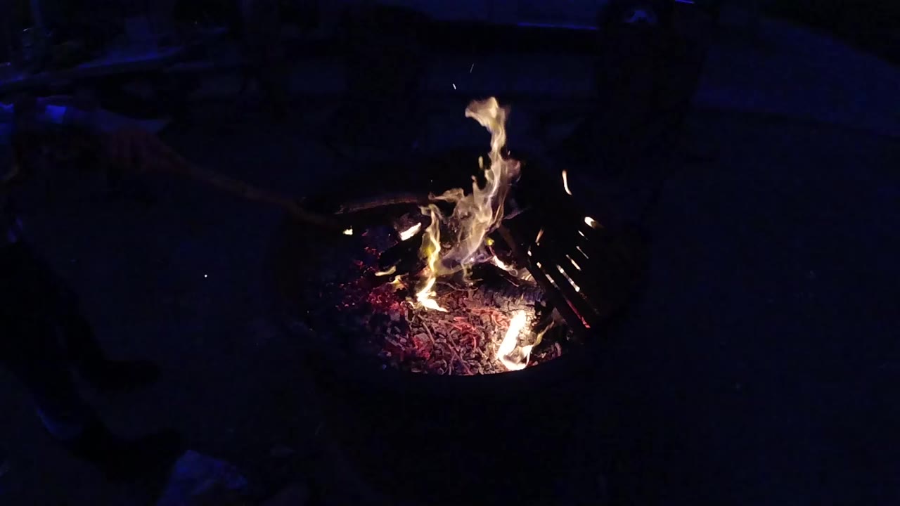 Metal fire pit with broad clustered yellow-white flames burning over red-purple embers at night
