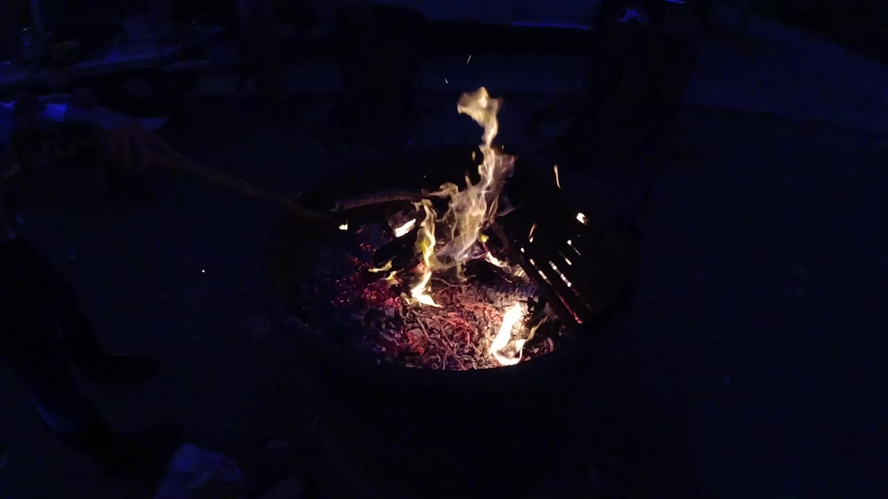 Overhead-angle view of the same metal fire pit at Trestles Beach California with flames stretching taller and more vertical than in the opening second, the ember bed still glowing red-purple against a near-black background; no people visible.