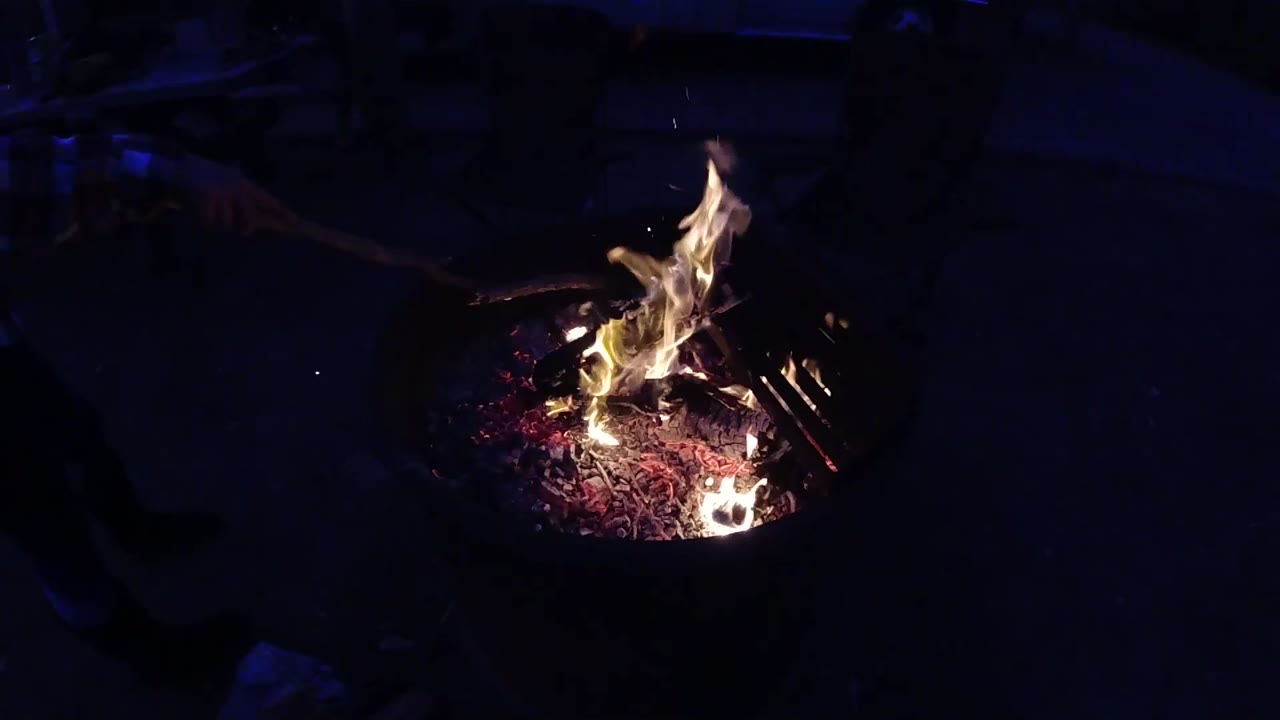 Overhead-angle view of the metal fire pit at Trestles Beach California with flames at similar height to the previous second; a dark linear element, possibly a structural edge or equipment, is faintly visible in the upper right corner; no people visible.