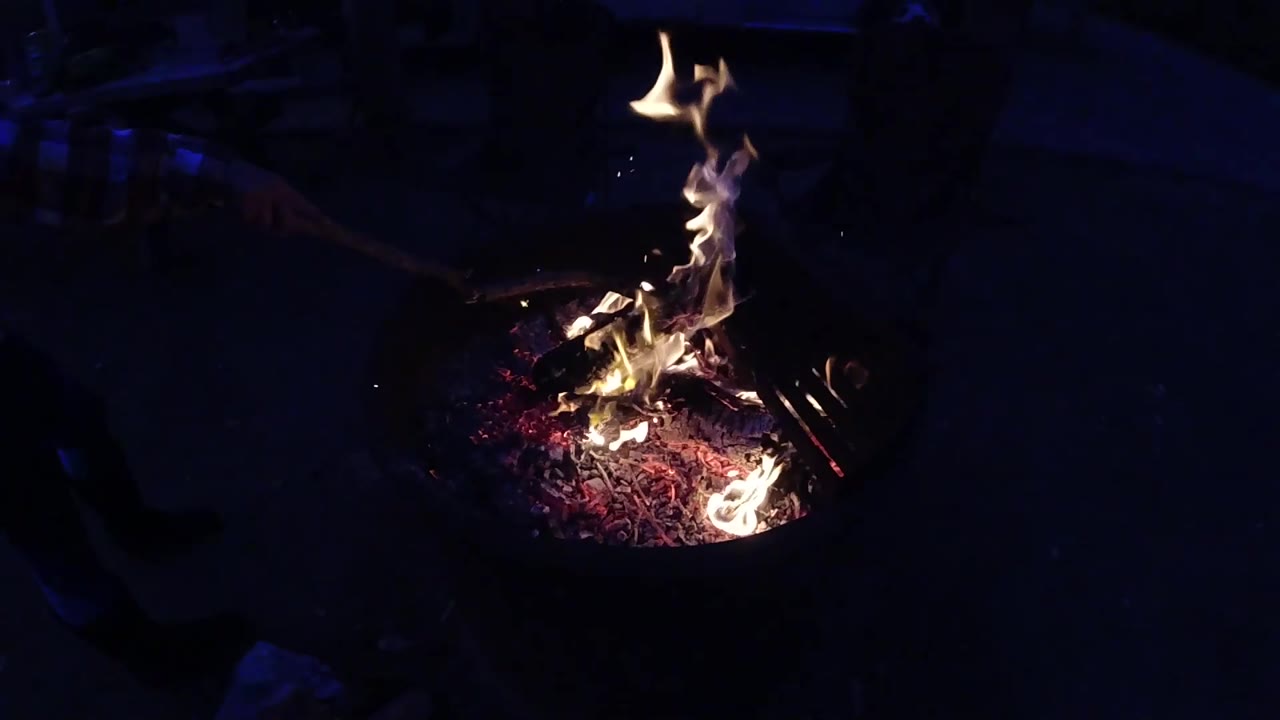 The fire in the metal pit at Trestles Beach California with the flame column leaning noticeably to the left; faint blue-tinted shapes, possibly gear or equipment, are visible at the left frame edge suggesting nearby objects; no people visible.