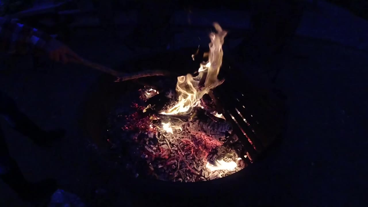 Elevated view of a round metal fire pit at night, flames active and rising from glowing red-orange embers and burning logs against a near-black background; no people visible; from the 2019 Trestles Beach California collection