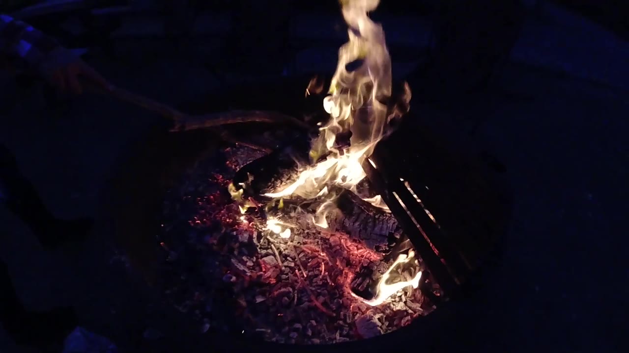 The right curved edge of the round metal fire pit becomes more prominent as the framing shifts slightly; orange-yellow flames and glowing embers are visible inside the pit; background is deep black; no people visible