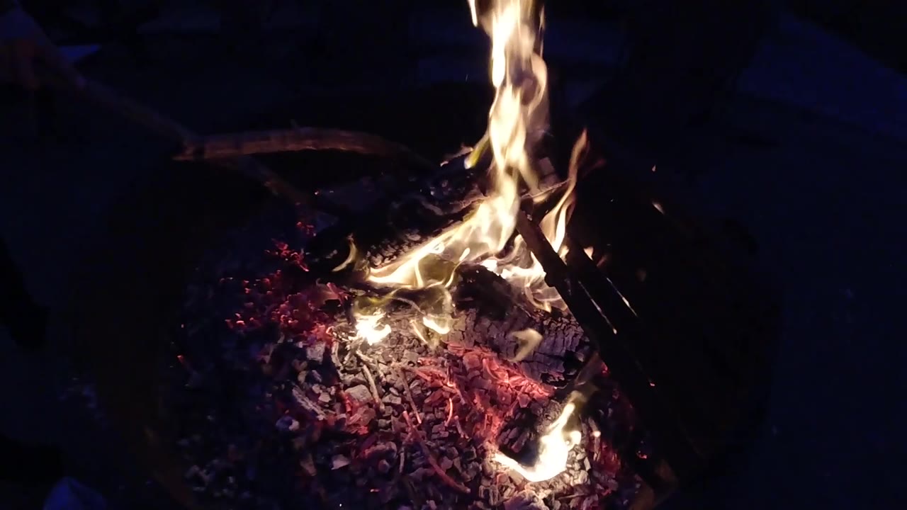 The camera has closed significant distance by three seconds; burning logs and deep red-orange embers now fill most of the frame, with orange-yellow flames rising and the fire pit's curved rim still visible at the right edge; no people visible