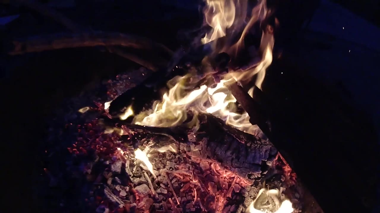 At two seconds, the campfire at Trestles Beach, California fills the frame with orange-yellow flames and glowing embers while the dark metal rim of the round fire pit enters the top edge of the composition; no people are visible