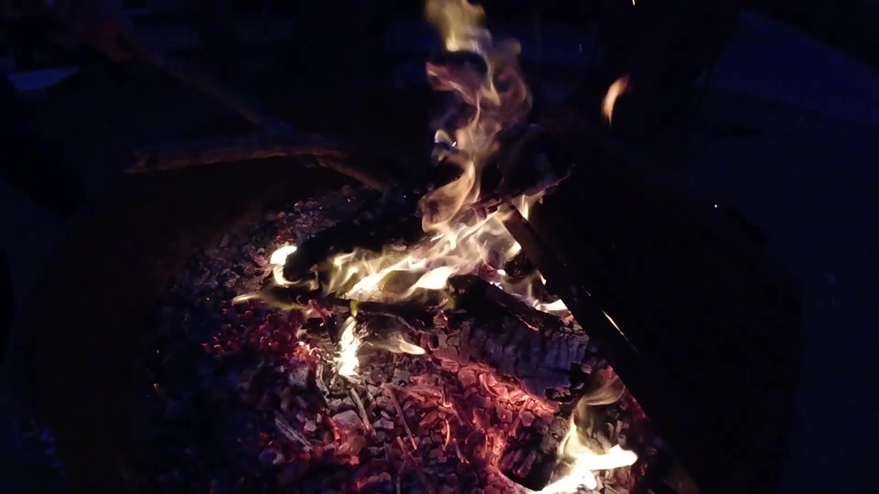 At three seconds the campfire at Trestles Beach, California drops from its earlier height to a low flicker, with a thin wispy flame rising from the embers and faint blue-purple ambient light appearing in the lower-left corner; no people are visible