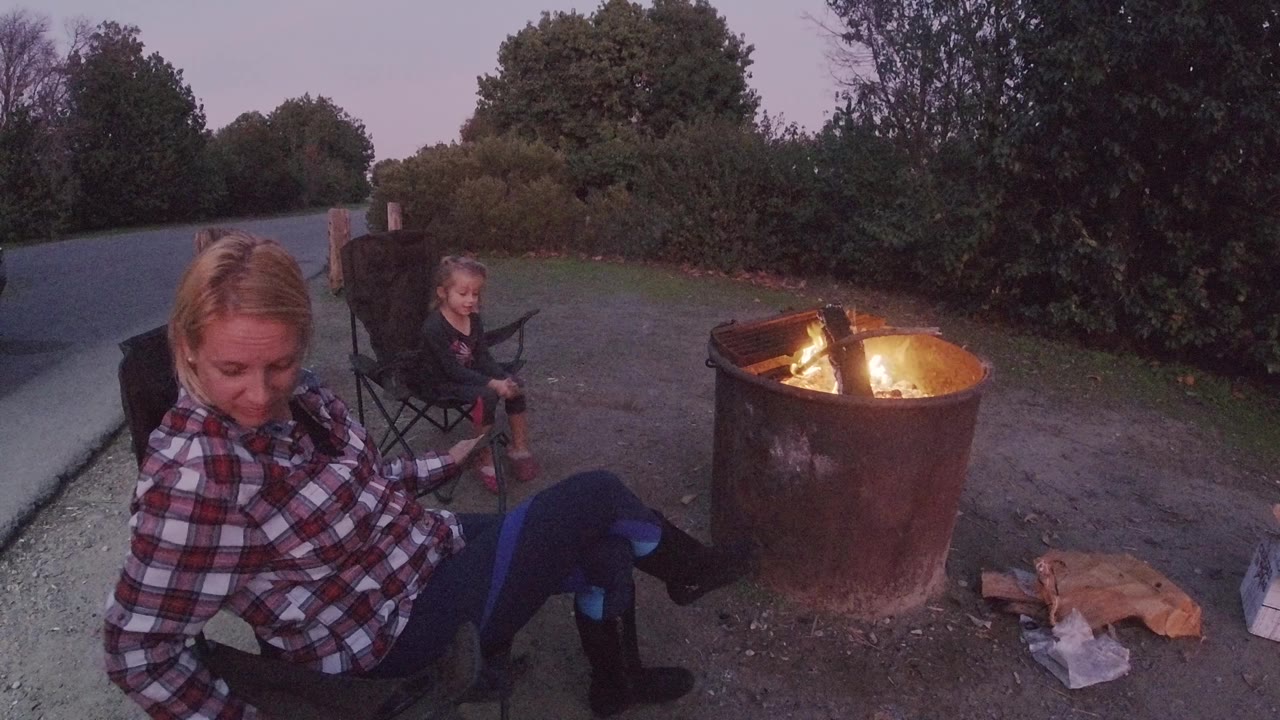 The woman looks downward from her chair as the child remains seated behind her and flames burn in a round metal fire ring at Trestles Beach, California.