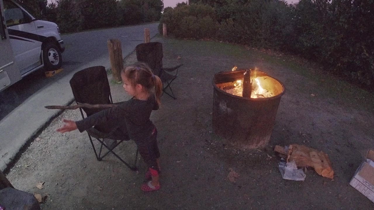 Young girl at Trestles Beach standing to the left of a large metal fire pit at dusk, holding a long stick extended toward the flames, with a folding camp chair and dark vehicle visible in the background, viewed from an elevated angle.