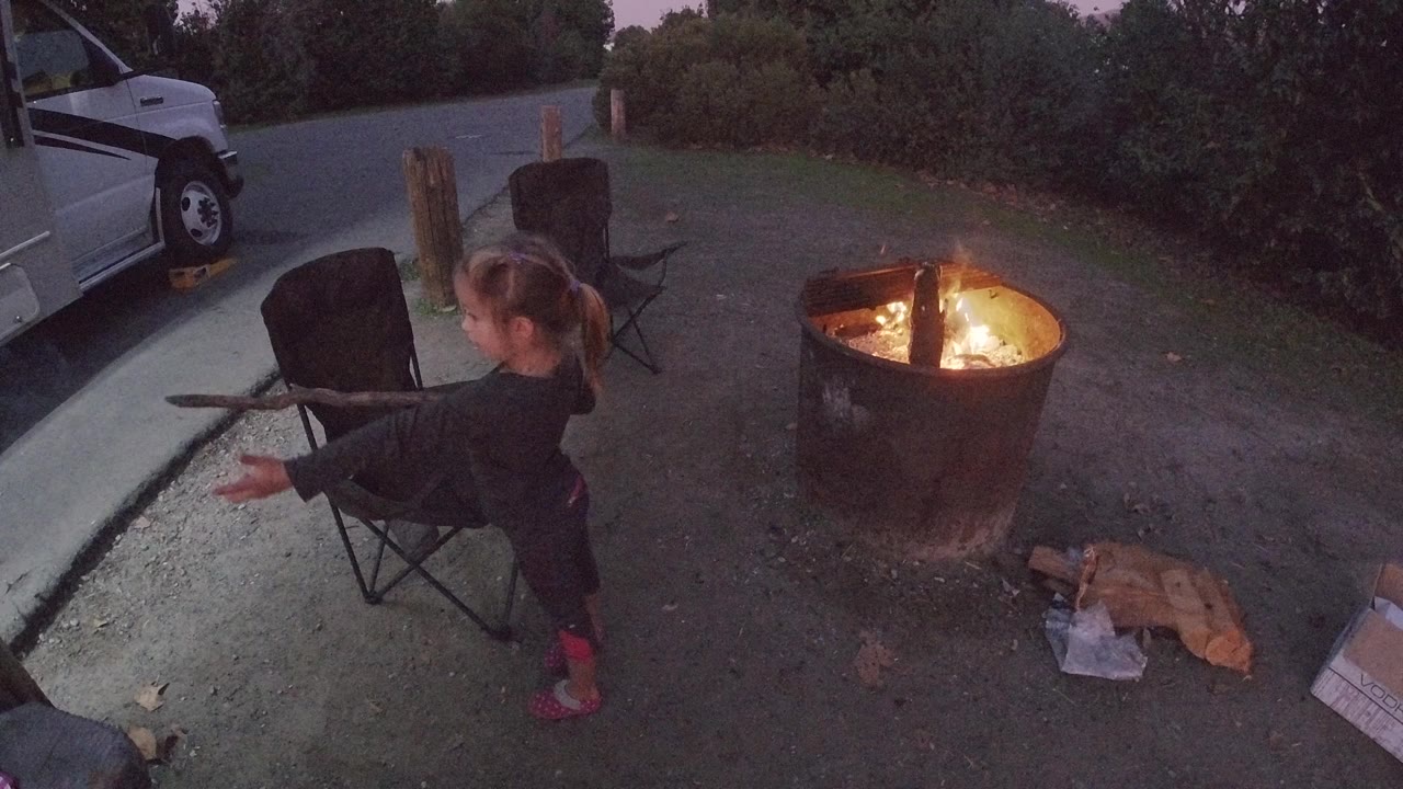 Young girl at Trestles Beach shifts her stick toward the fire pit at one second in, her body turned slightly more toward the flames compared to the opening frame, with the folding chair and vehicle unchanged in the background.