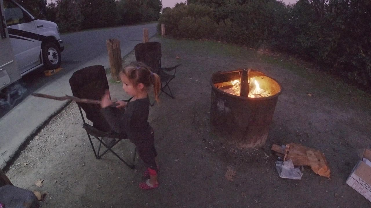 Young girl at Trestles Beach holds her stick in a slightly different position from frame 1, with the fire pit flames at right and the camp chair at left, the dusk sky noticeably cooler in tone than the warm fire glow below.