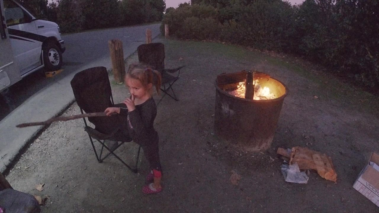 Young girl in black clothing and red shoes stands with a stick near a fire pit at dusk, viewed from an elevated angle.