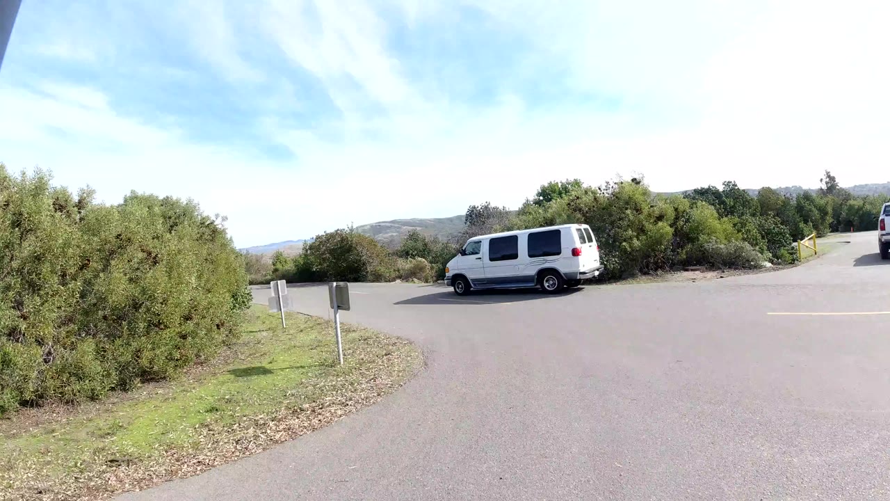 A San Clemente, California road bend shows shrubs, grass, small roadside signs, a white van, distant hills, and bright midday sky, with no visible people.