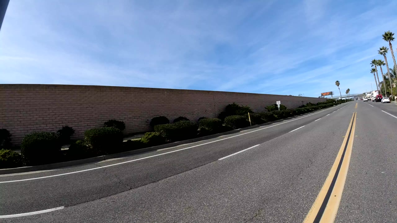 Street-level view of a San Clemente road beside a long block wall, with double yellow lines, distant cars, palm trees, and no visible people.