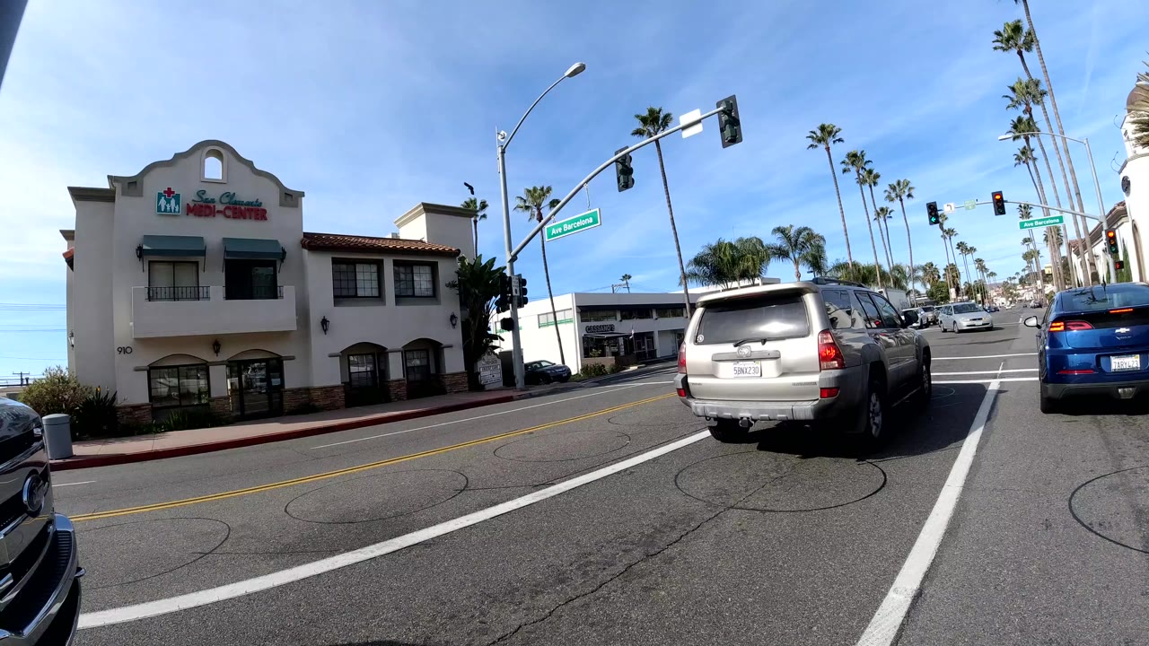 Wide street-level view in San Clemente with a silver SUV and blue car near a traffic light, a two-story commercial building on the left, palm trees, and no clearly visible people.