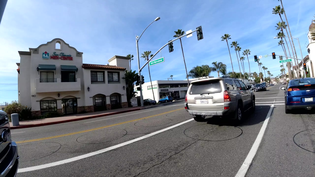 Similar San Clemente intersection view with vehicles closer to the camera, red traffic signals overhead, a low storefront across the street, and no clearly visible people.